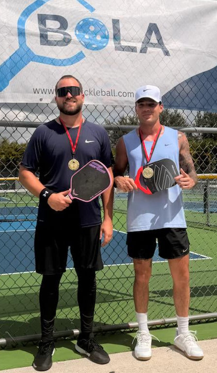 Two men on a tennis court holding paddle sports equipment with a BOLA banner in the background.